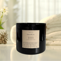 A black 20oz jar candle with a label that reads 'SANCEO 1111', placed on a white surface with a white towel and some foliage in the background.